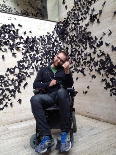 Man in a wheelchair, who is wearing glasses and has a beard. He’s enthusiastically giving a thumbs-up with his right hand, showcasing a positive spirit. The background is a captivating