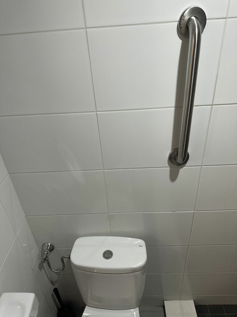 Baño con inodoro blanco, A la derecha del inodoro, hay una barra de seguridad metálica fijada a la pared, que está cubierta con azulejos blancos.