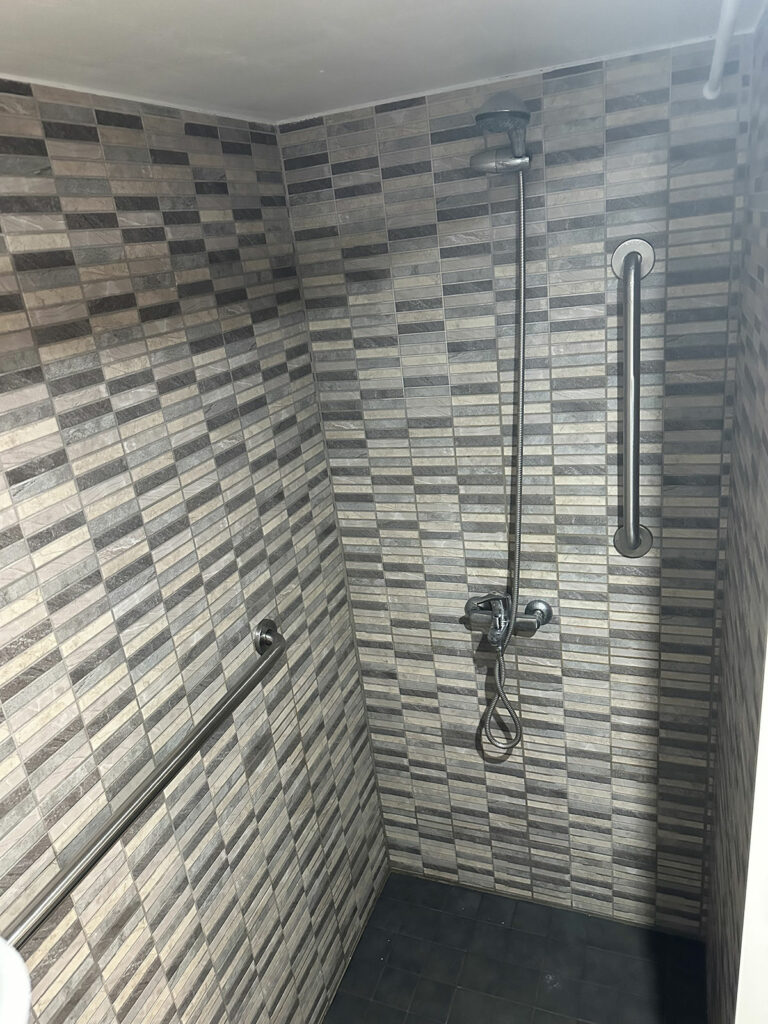 Área de ducha con paredes y suelo de azulejos rectangulares. Suelo en tonos grises, aspecto texturizado. Hay una barra de seguridad horizontal fijada a la pared izquierda, y una barra de seguridad vertical en la pared derecha. Un cabezal de ducha está montado en la pared, con una manguera colgando y un soporte circular para el cabezal de ducha.