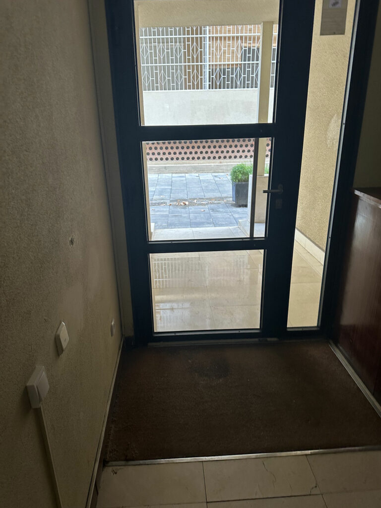 Interior de edificio con puerta de vidrio y marco metálico de color oscuro en tres partes que facilita la visibilidad. Incluye barra vertical que facilita el agarre de la puerta y felpudo insertado al suelo con contraste de color marrón. Las paredes interiores son de color claro, hay accionadores visibles en la pared izquierda a una altura de 90 centímetros.