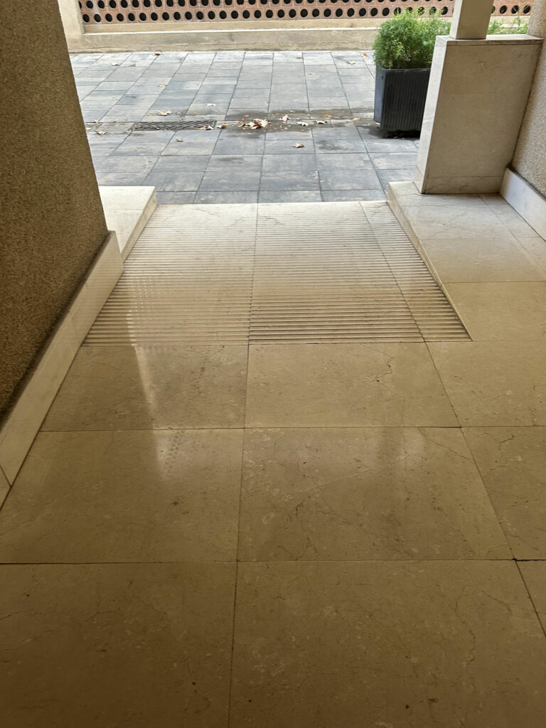 Vista de superficie con rampa que lleva hacia el exterior. Hoy franjas de pavimento podo táctil en la superficie, guías para personas con discapacidad visual. El sendero está flanqueado por paredes a ambos lados, y conduce a un área exterior con una superficie pavimentada.
