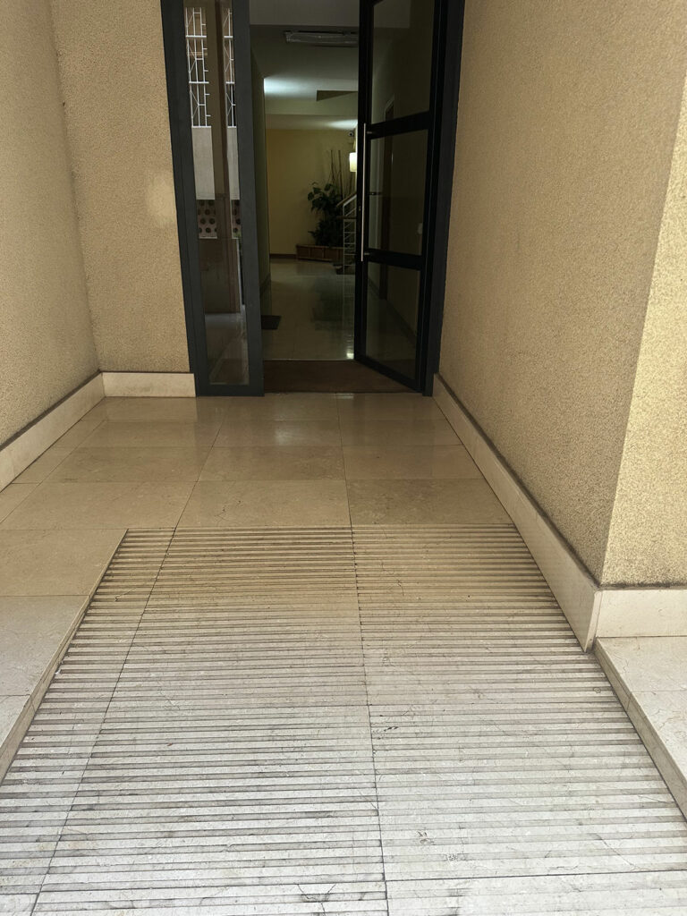 Vista desde el exterior hacia la entrada del edificio residencial. El cambio de nivel en la entrada se salva con una rampa, con la superficie señalizada con pavimento táctil para peatones. La entrada cuenta con una puerta de vidrio con control automático de apertura.