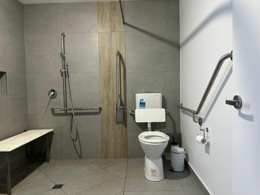 Baño con inodoro blanco. Hay barras de apoyo metálicas, en ambos lados del inodoro. En la parte izquierda, hay un asiento abatible para ducha y un grifo con una ducha de mano flexible adaptado a una barra vertical. El suelo y las paredes están revestidos con azulejos de color gris claro.