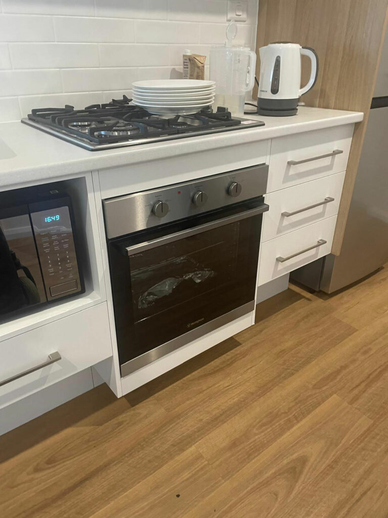 Cocina accesible con encimera blanca y estufa de gas a la izquierda. Debajo, horno y microondas a una altura adecuada para personas en silla de ruedas. Suelo de madera clara y cajones blancos con tiradores fáciles de usar. Espacio suficiente debajo de los mostradores para una movilidad cómoda con ayudas para desplazarse.