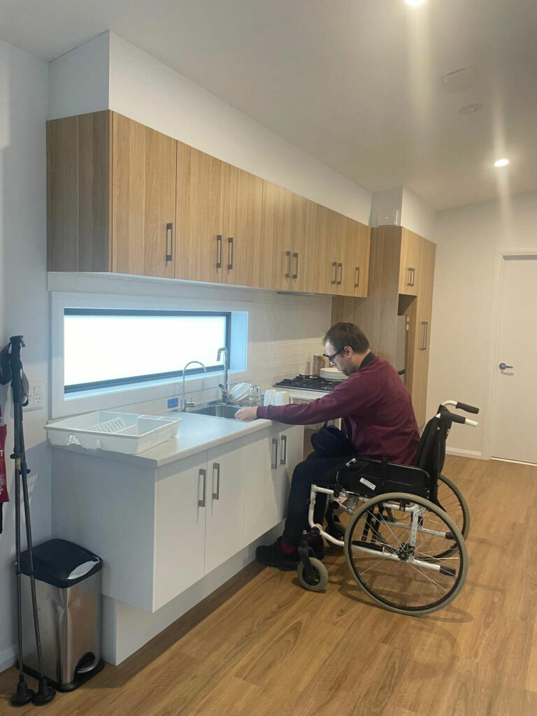 Hombre en silla de ruedas lavando un plato en una cocina accesible, con armarios a la altura adecuada nevera, cocina y fregadero ubicados en línea y un diseño espacioso que facilita el movimiento. El suelo es de madera clara y una ventana sobre el fregadero aporta buena iluminación.