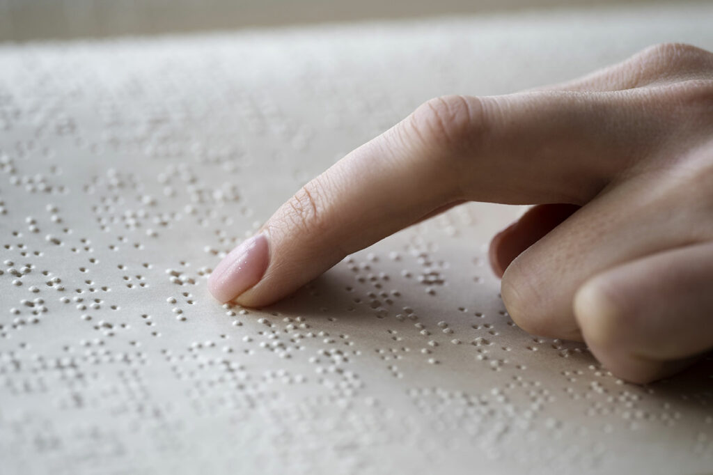 La imagen muestra un primer plano del dedo de una persona tocando una página con texto en braille. Los puntos del braille están elevados en la superficie del papel, lo que permite leerlos mediante el tacto. El dedo está posicionado en el lado derecho de la imagen, descansando suavemente sobre el braille.