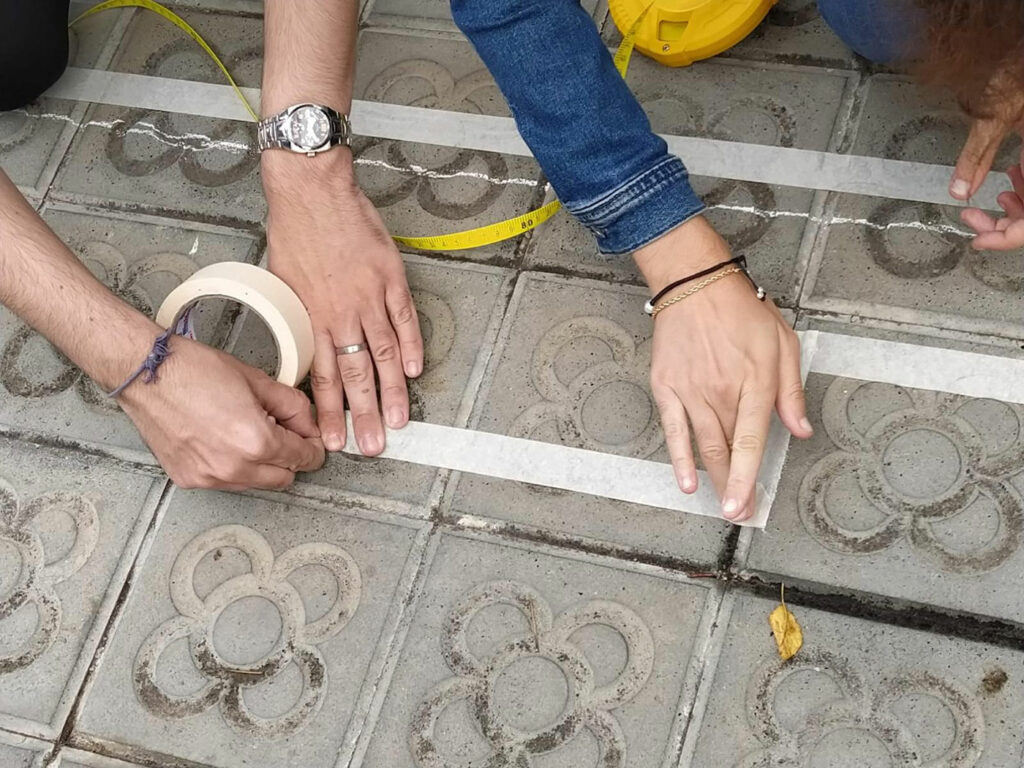 Manos trabajando con un rollo de cinta adhesiva sobre una superficie en zona exterior para simular el plano en escala real de una casa accesible, esta actividad es realizada por personas de la comunidad.