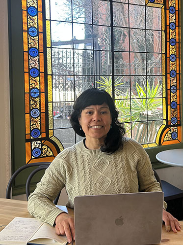Woman with long black hair, dressed in a light-colored sweater, seated at a wooden table with a laptop in front of them. Behind them, a large, colorful stained glass window featuring intricate designs  reates a vibrant backdrop. This indoor setting beautifully complements the scene.